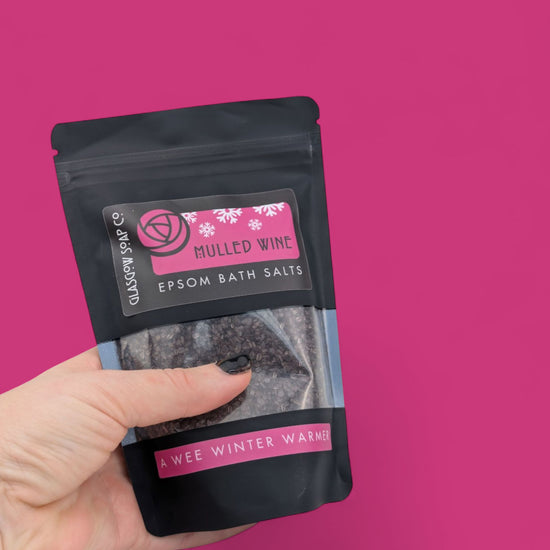 Hand holding a package of mulled wine Epsom bath salts against a pink background