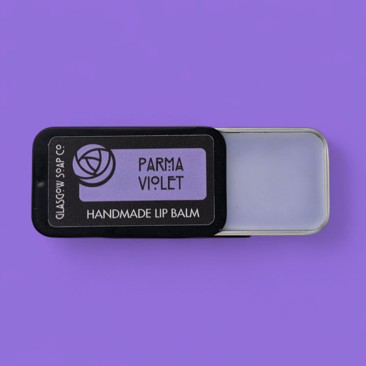 Handmade lip balm in a black tin labeled 'Parma Violet' on a purple background