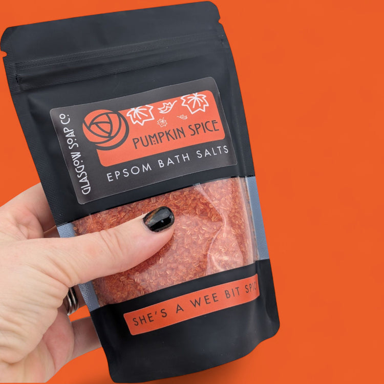 Hand holding a package of pumpkin spice Epsom bath salts against an orange background