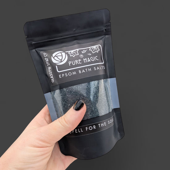 Hand holding a black package of Epsom bath salts with 'Pure Magic' label on a dark background
