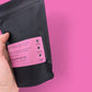 Black package with pink label held by a hand against a pink background