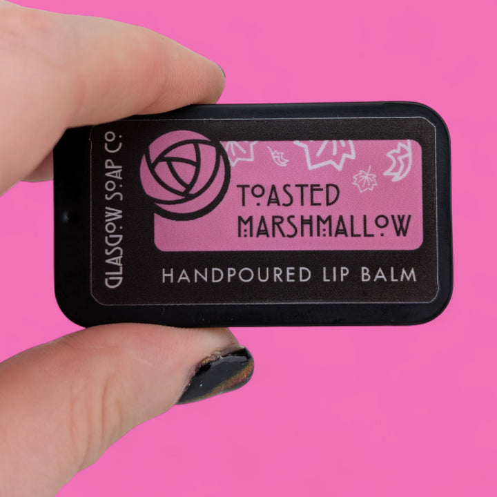 Hand holding a lip balm labeled 'Toasted Marshmallow' against a pink background